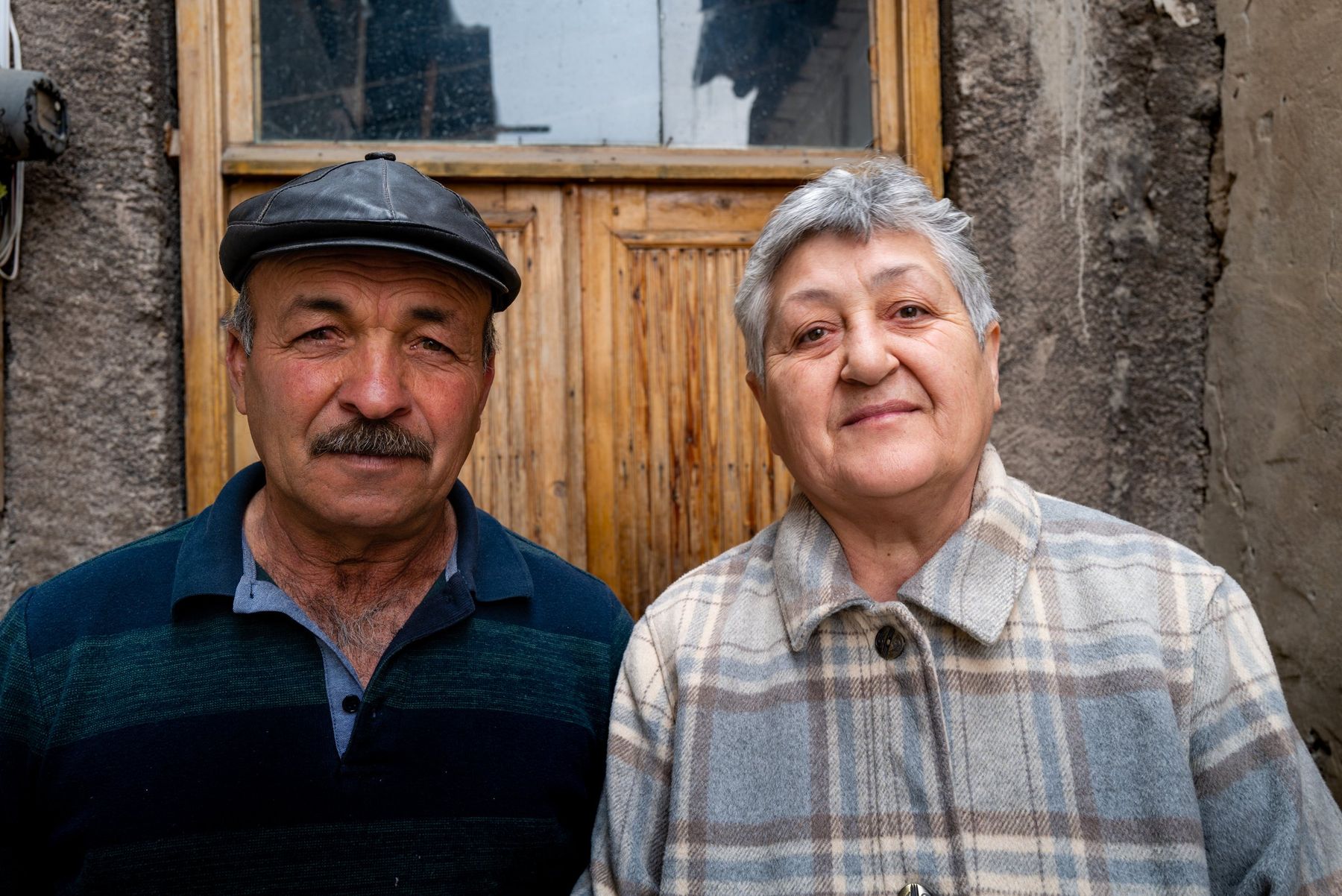 An elderly couple stand together in front of a wooden door