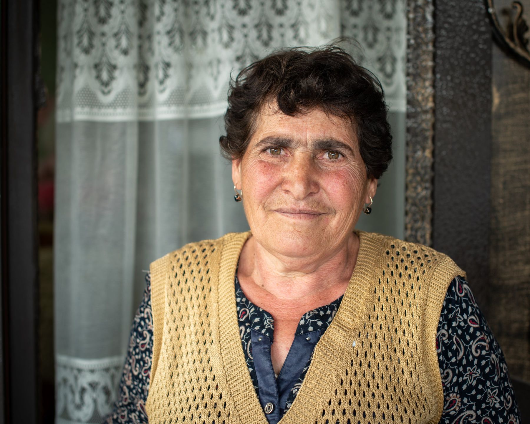 A woman in a knitted waistcoat stands in a doorway, framed by a lace curtain