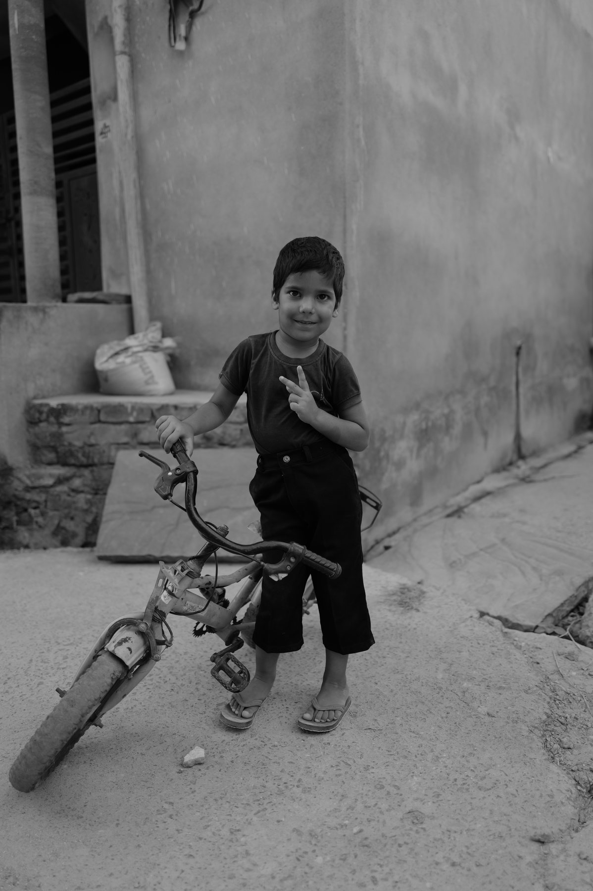 A small boy stands beside a bicycle in a lane, looking at the camera