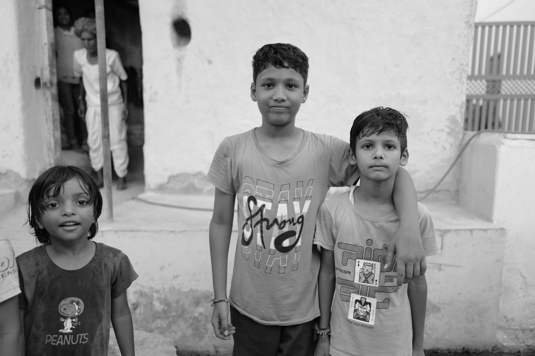 Three children stand in front of a house, two of them grinning