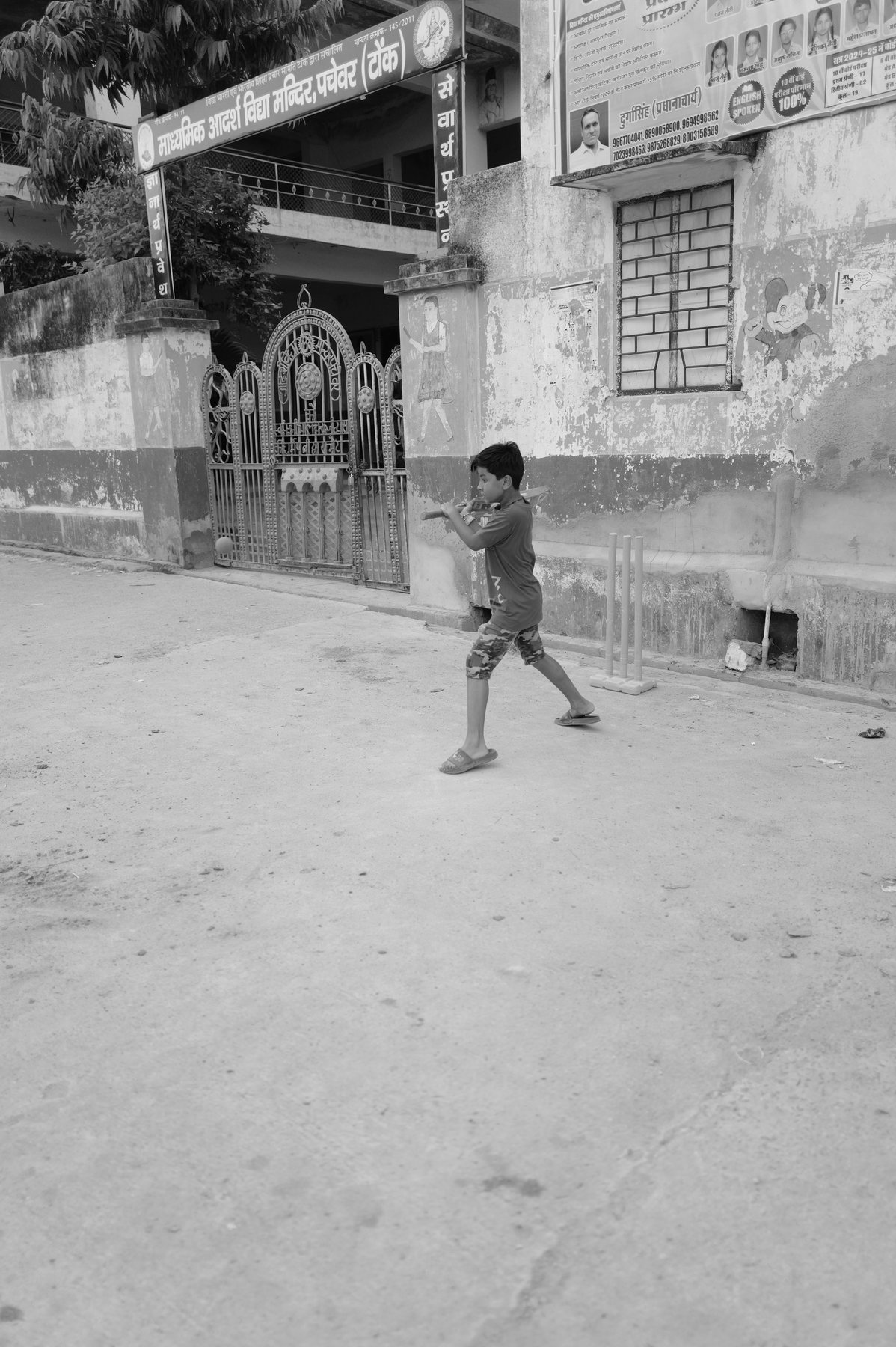 A boy swings a cricket bat in a dusty lane in front of a decorated gate