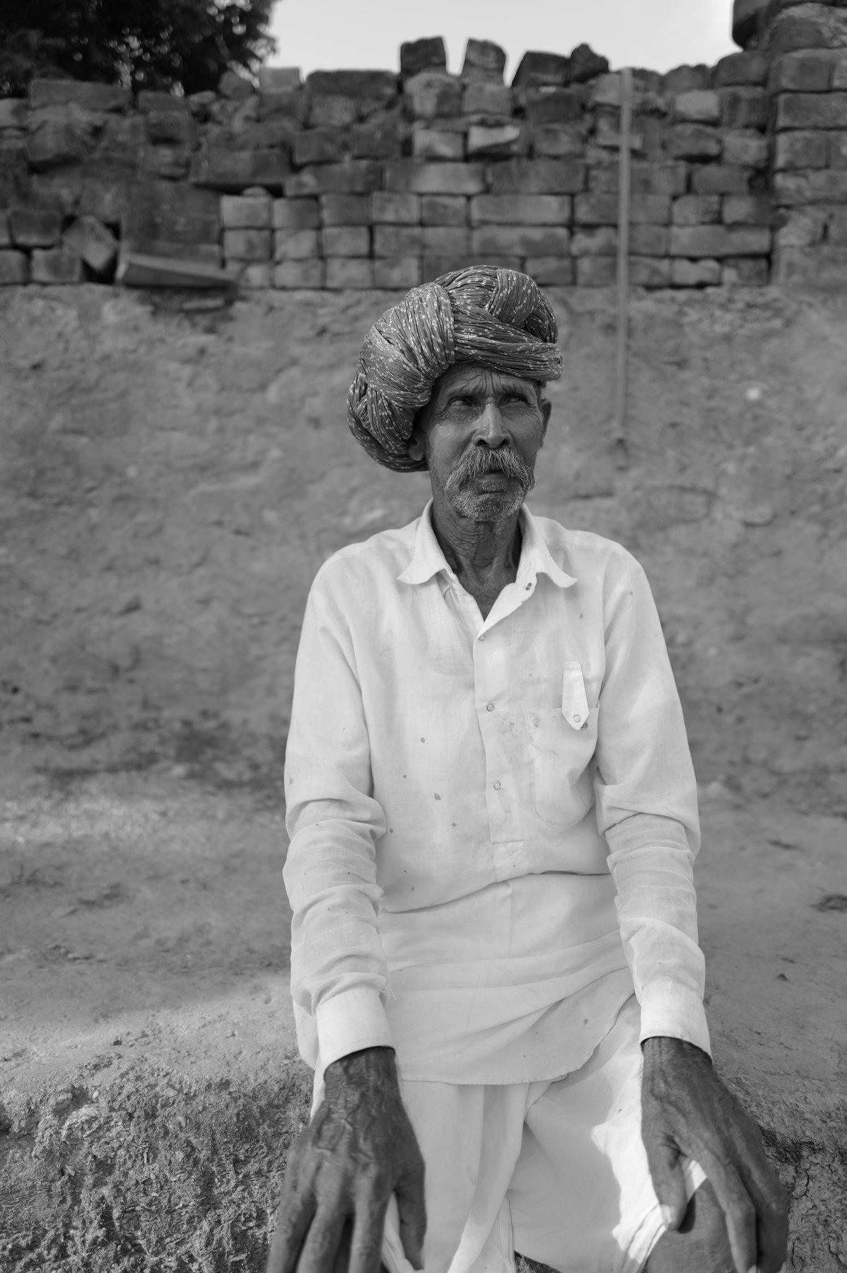An elderly man in a large patterned turban and white shirt stands before a brick wall