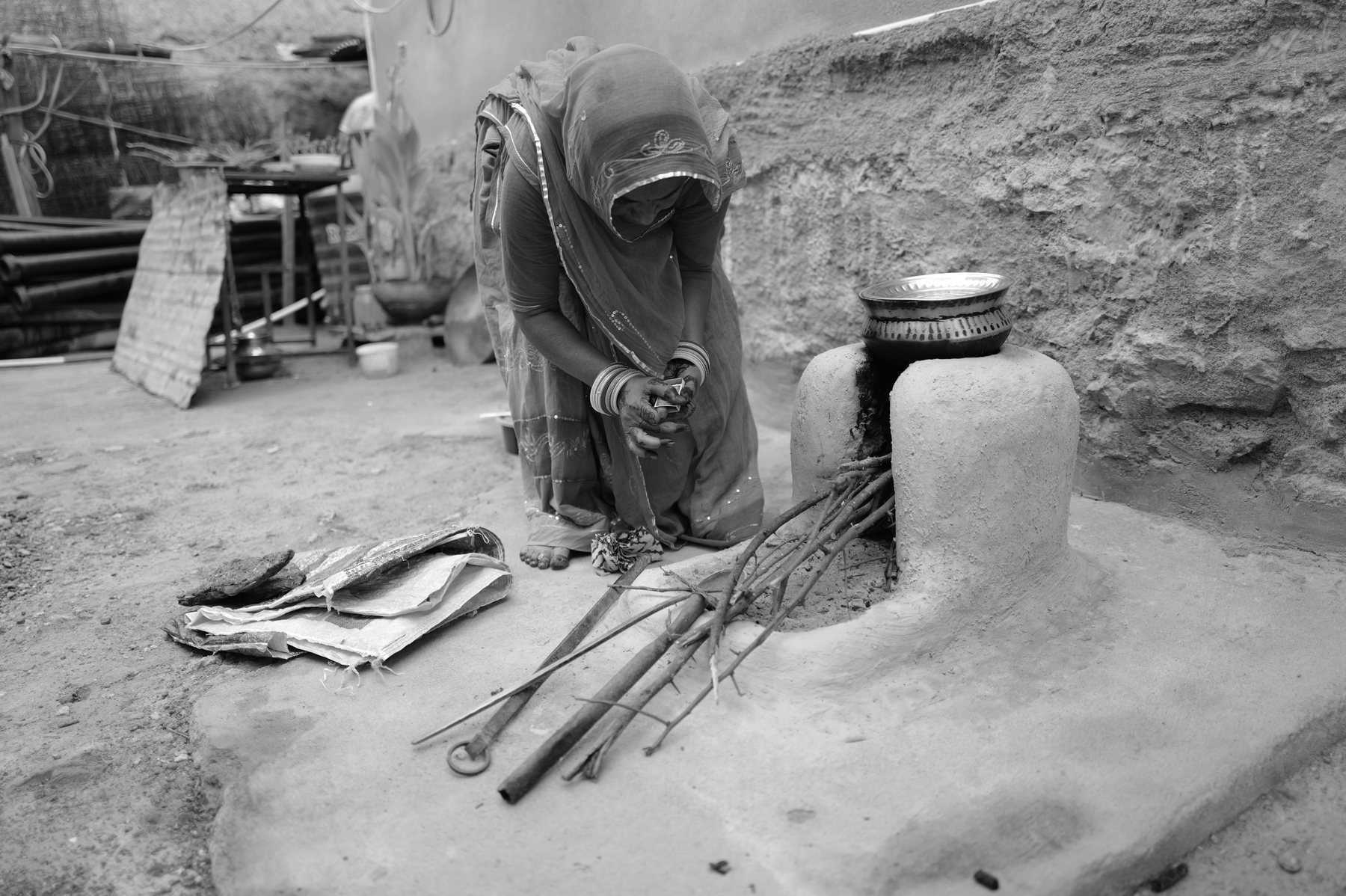 A woman in a headscarf kneels at the clay stove as the fire takes