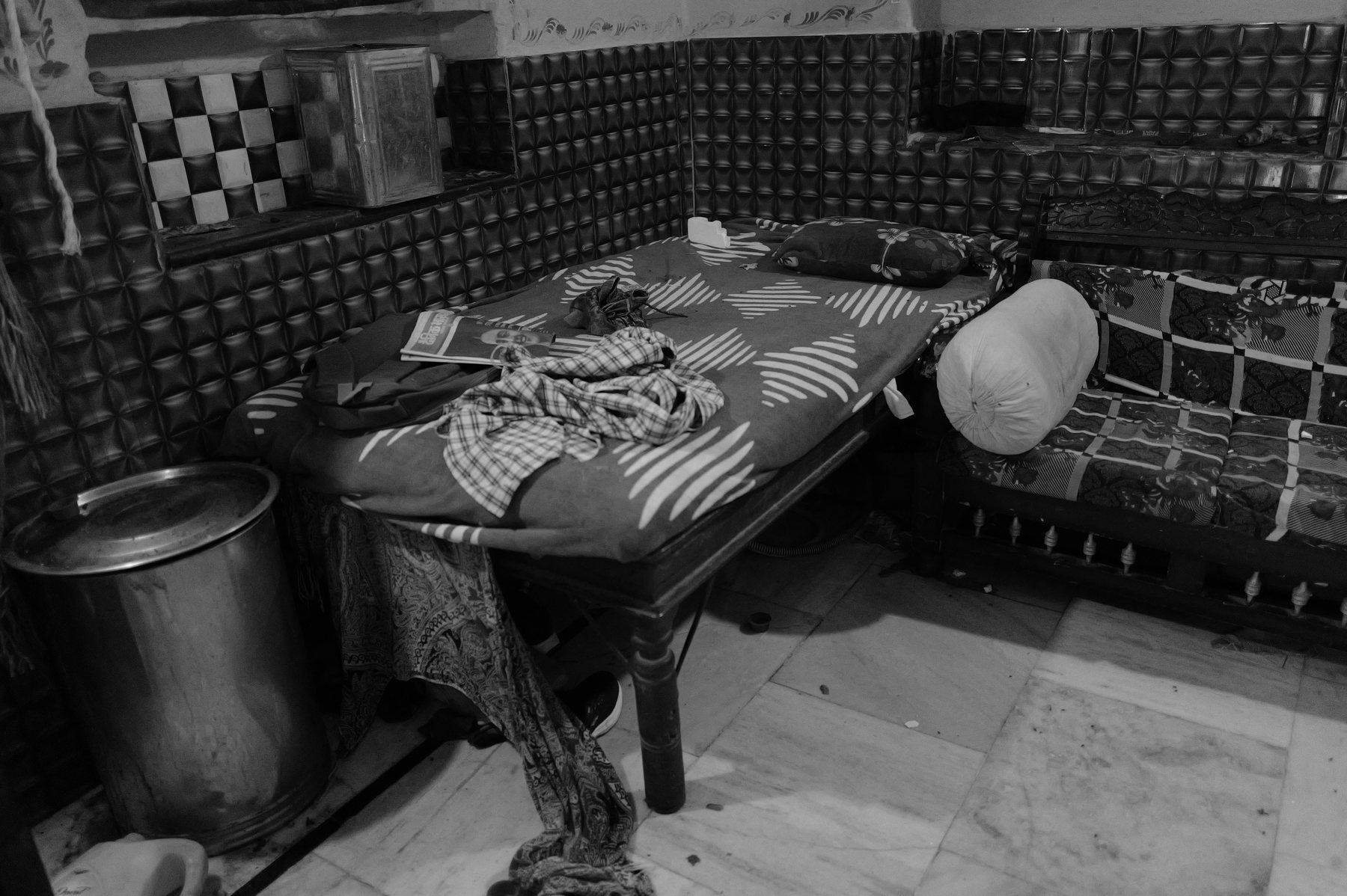 A bed with a patterned cover against a tiled wall, a pot beside it
