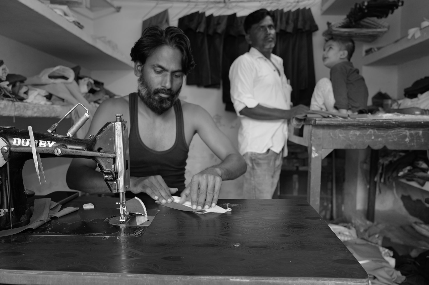 A young tailor works at a treadle sewing machine while an older man stands behind him