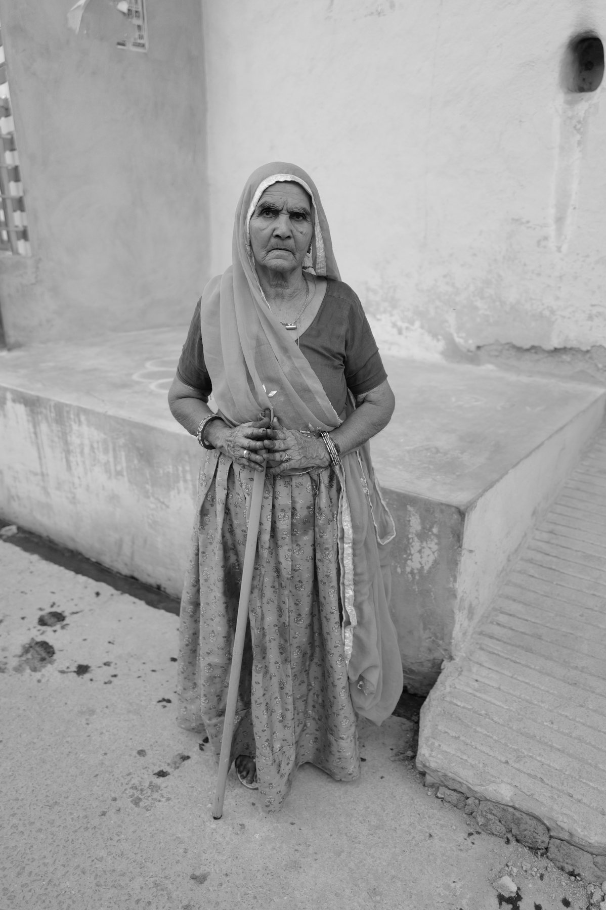 The same woman stands full length with a walking stick beside a whitewashed step