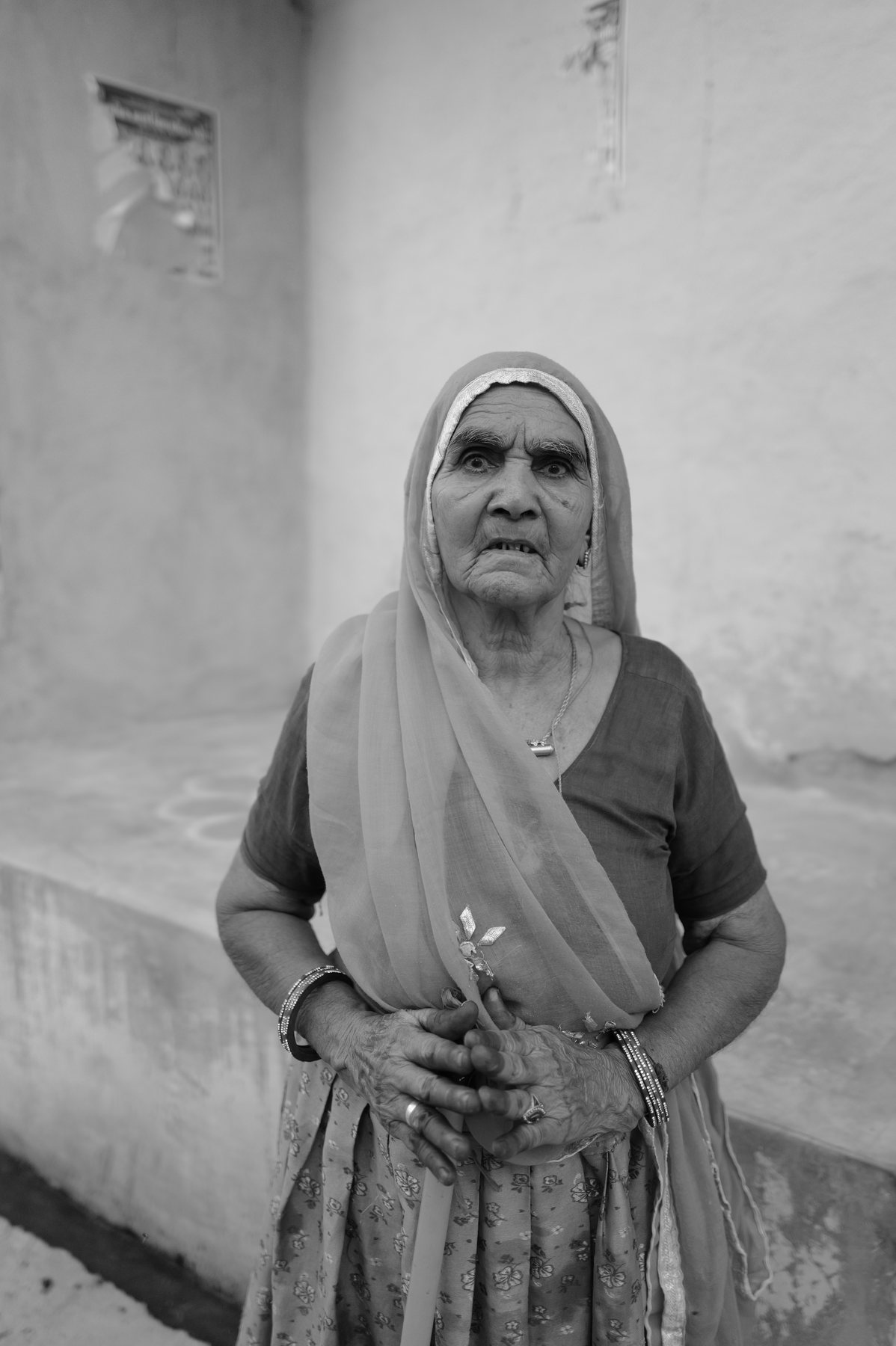 An elderly woman in a headscarf stands in a doorway, hands folded